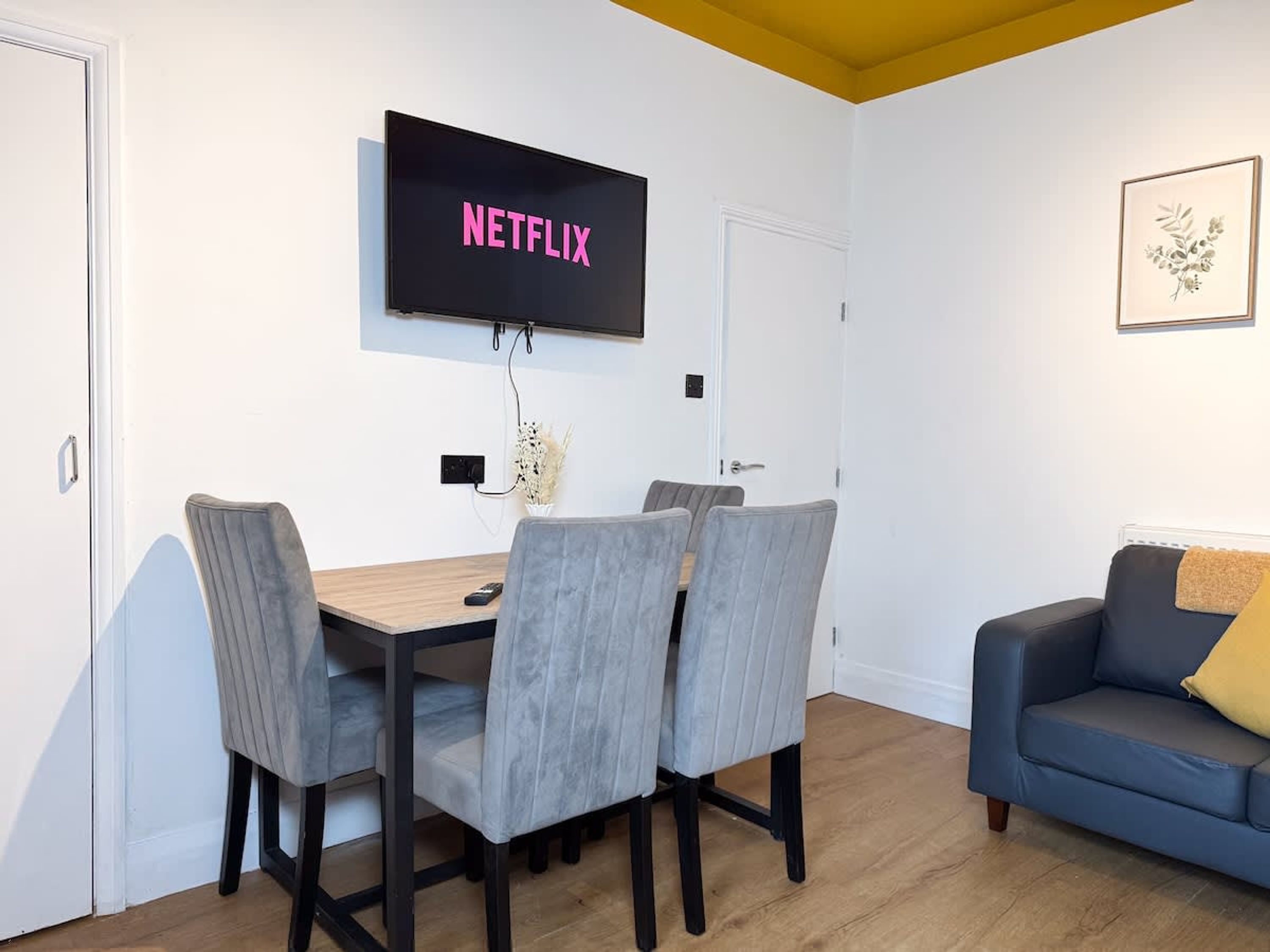 Un salon comprenant une table à manger, quatre chaises, un canapé et un téléviseur fixé au mur, sur lequel apparaît le logo de Netflix.