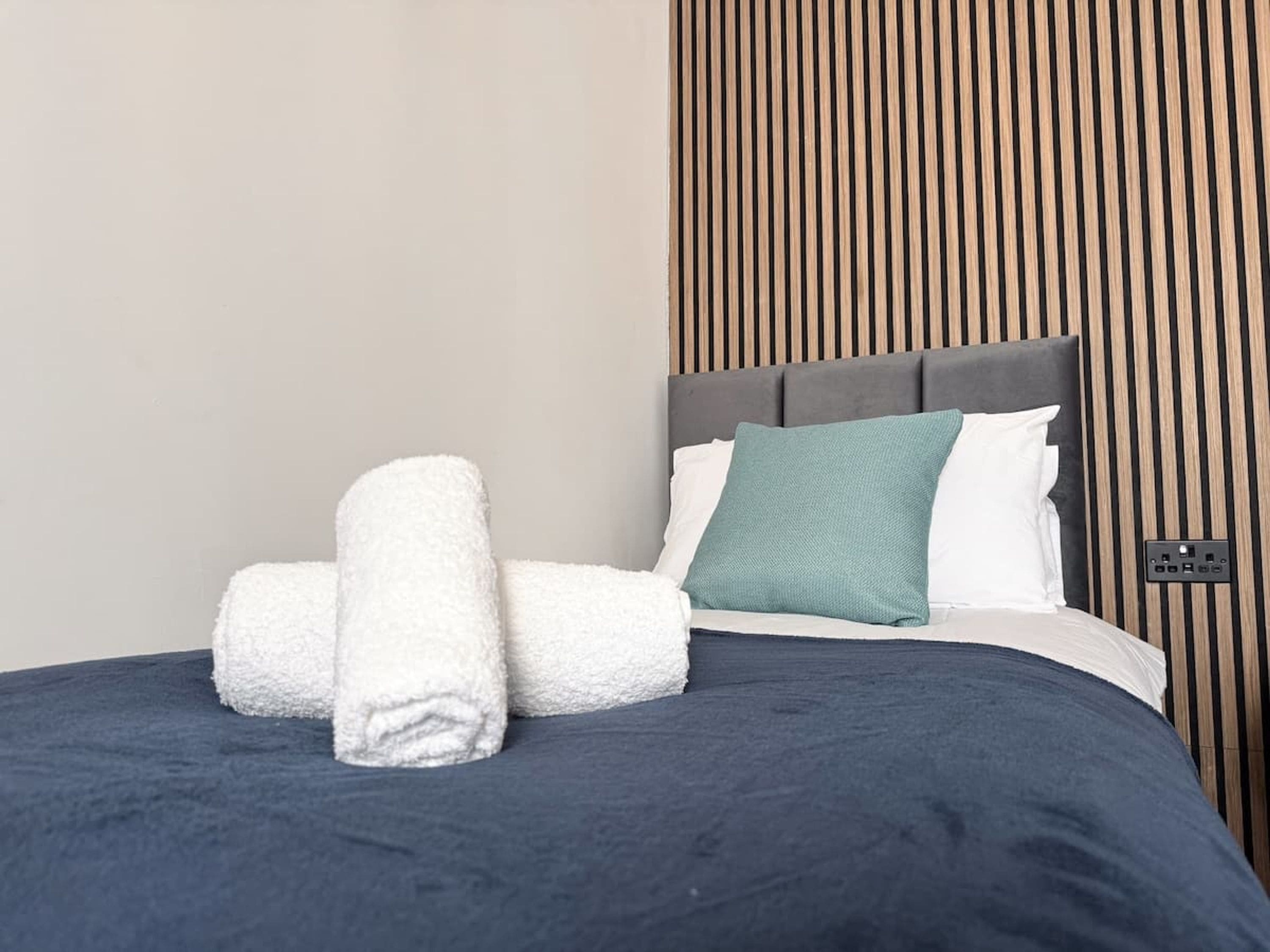 A neatly made bed with a grey headboard, white and teal pillows, and two rolled white towels.