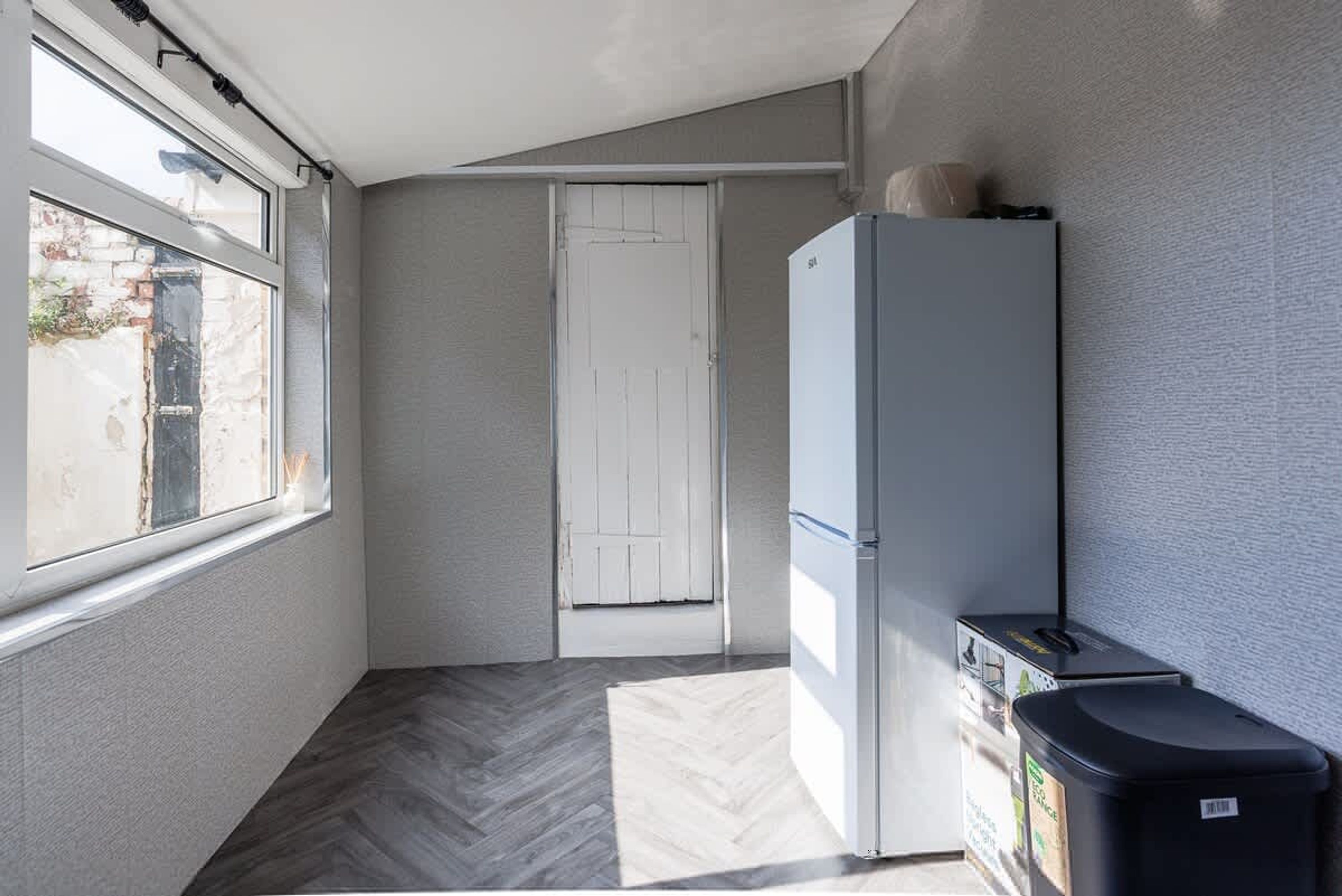 A narrow hallway with a white door, a white refrigerator, and a trash bin.