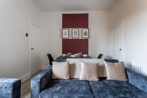 Ein modernes Wohnzimmer mit einem blauen Sofa, einem Esstisch für vier Personen und einer roten Akzentwand.