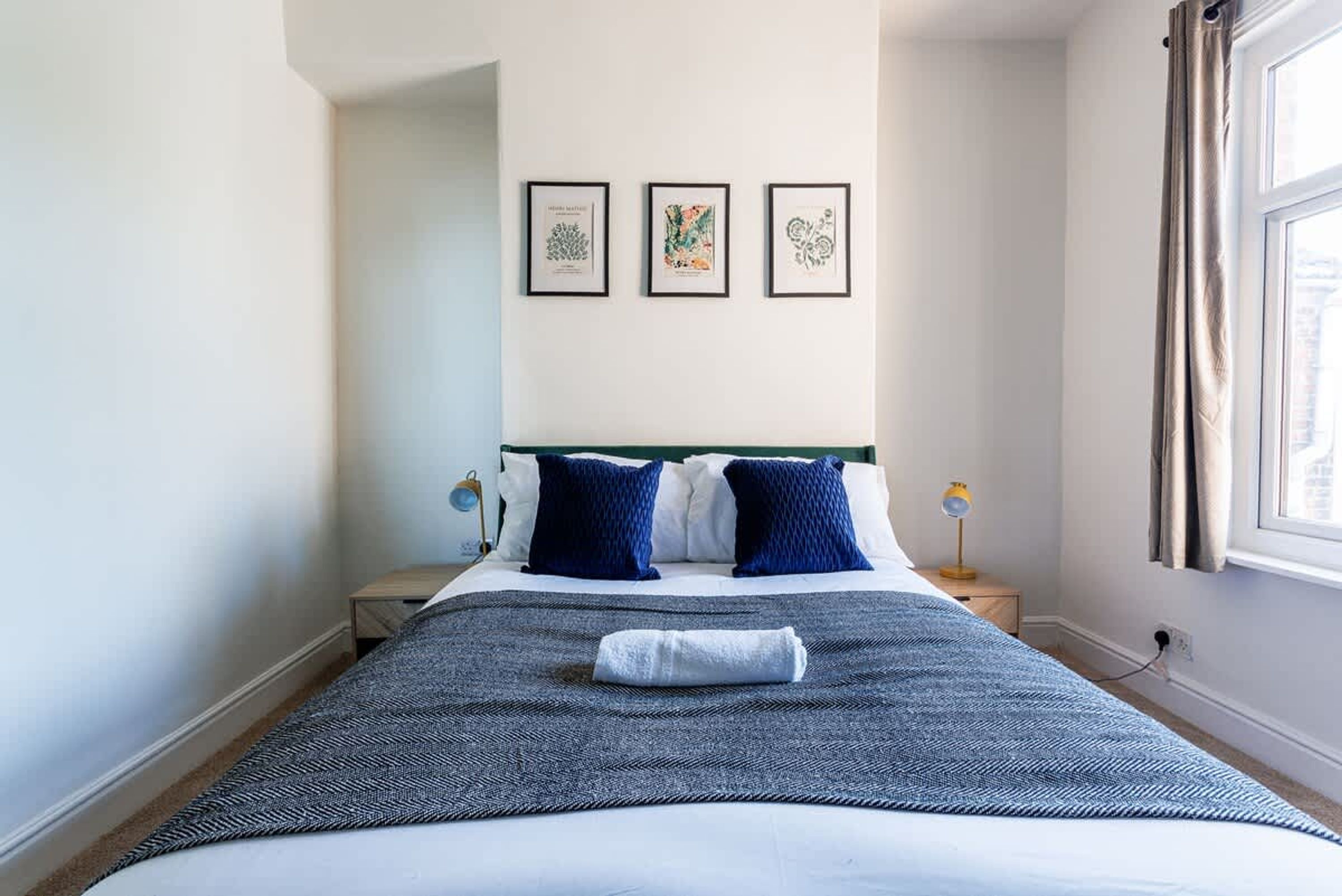 Un lit bien fait, recouvert d’une couvre-lit à motifs bleus et blancs, avec deux oreillers bleus et une serviette pliée posée sur le lit. Trois tableaux encadrés sont accrochés au mur au-dessus du lit.