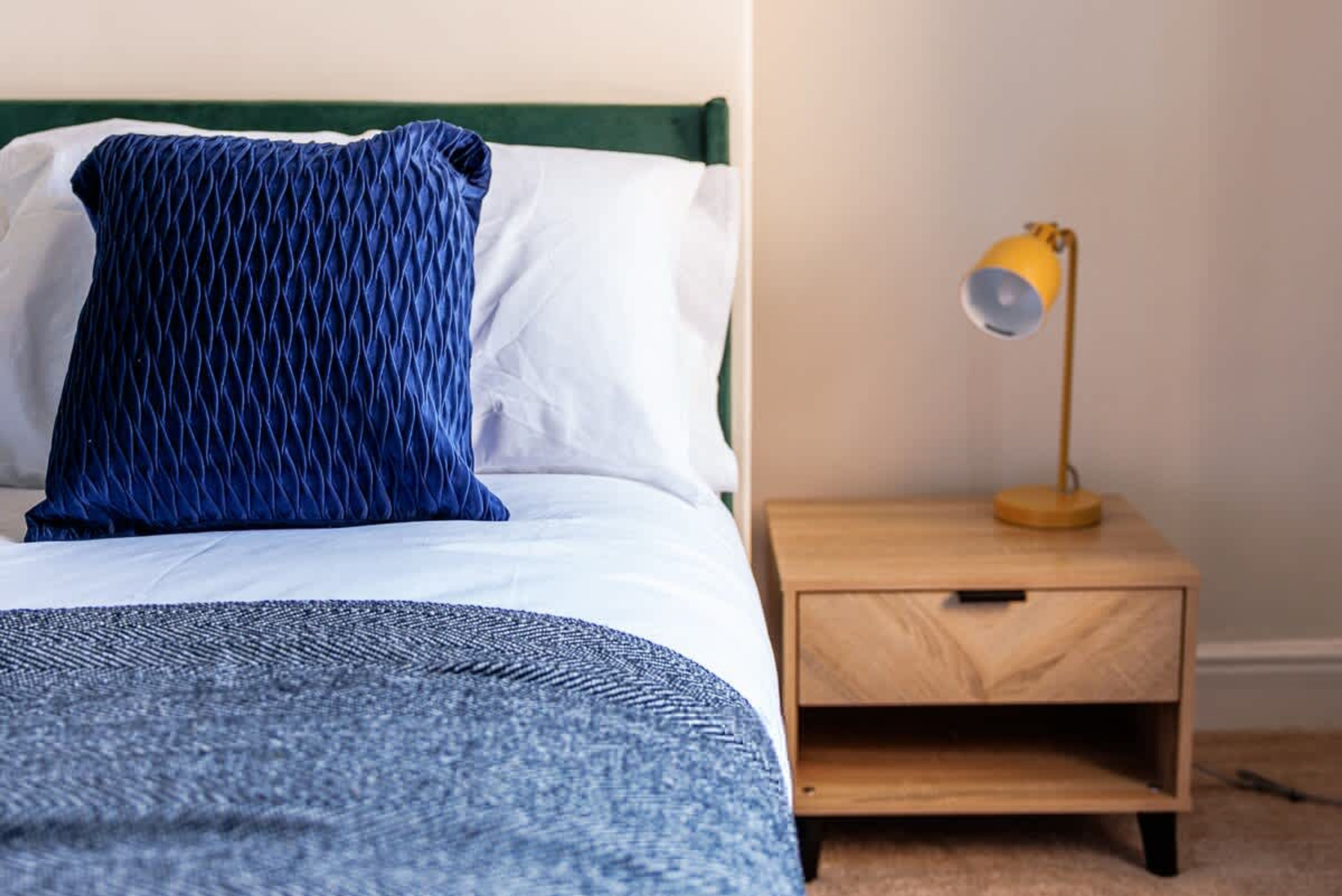 Un lit avec un oreiller à motifs bleus et un oreiller blanc, une couverture bleue, une table de chevet en bois avec une lampe et un tiroir en bois.