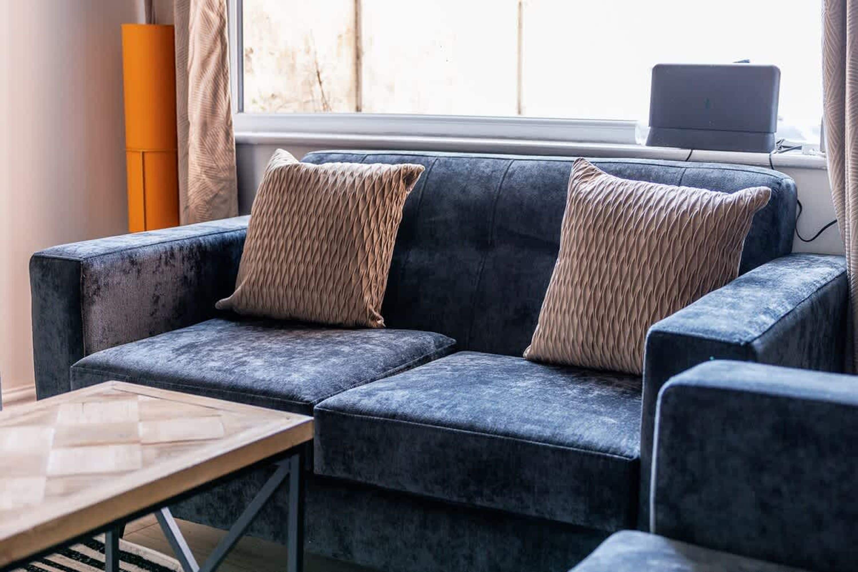 A modern living room with a dark blue sofa, two matching cushions, a wooden coffee table, and a laptop on a windowsill.