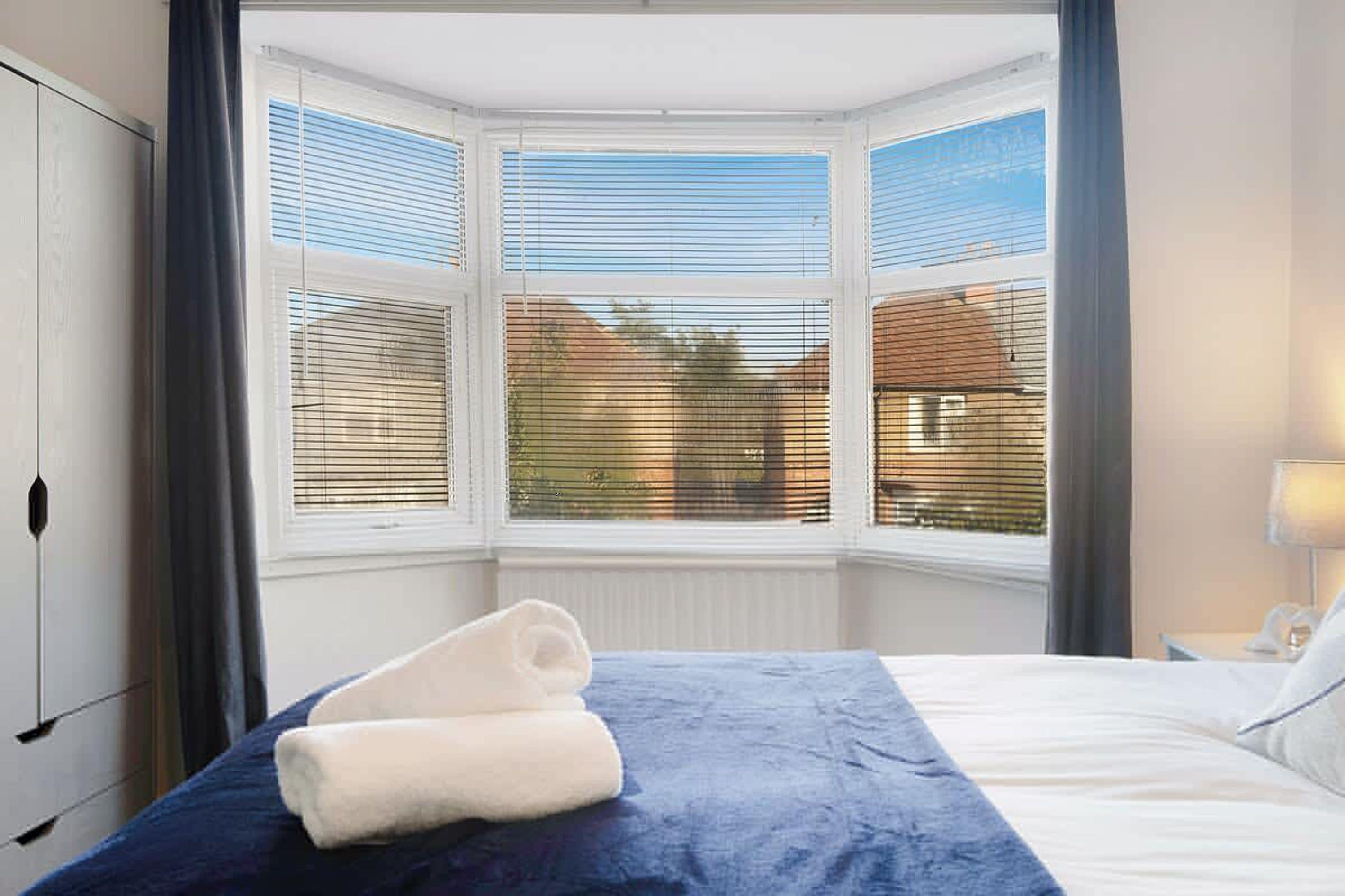 Habitación de hotel con cama, ventana con persianas, toalla sobre la cama y lámpara en la mesita de noche.