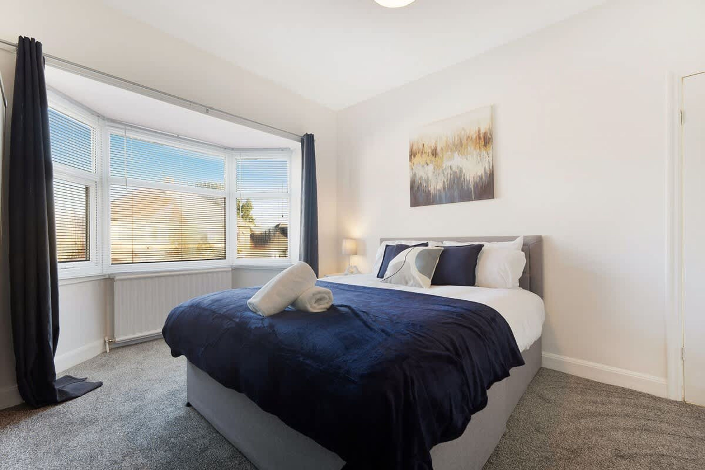 Habitación de hotel con una cama, una ventana con persianas, un cuadro en la pared y piso alfombrado.
