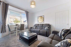 Een moderne woonkamer met een bank, fauteuils, een salontafel en een groot raam met gordijnen.