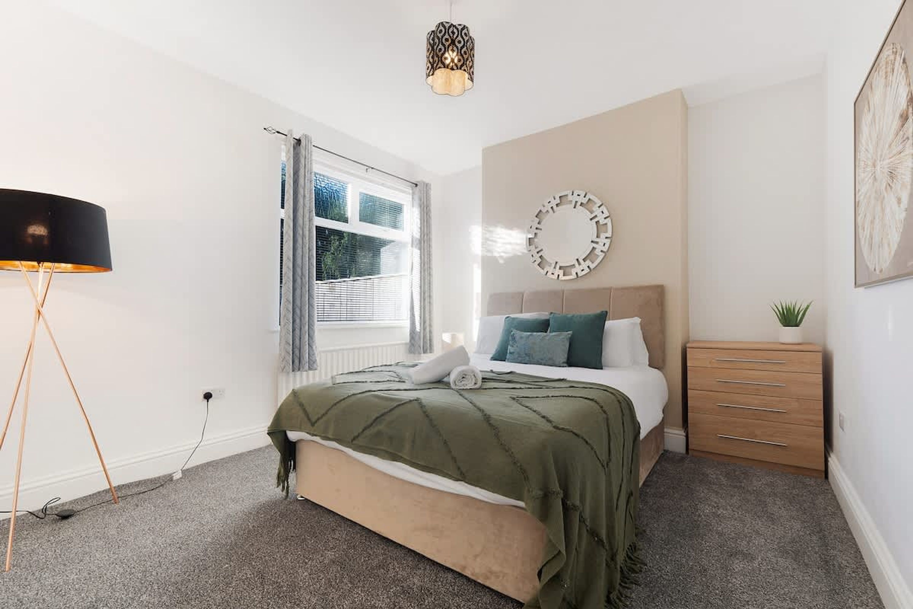 Une chambre à coucher comprenant un lit, une table de chevet, une commode, une fenêtre avec des rideaux et un miroir fixé au mur.