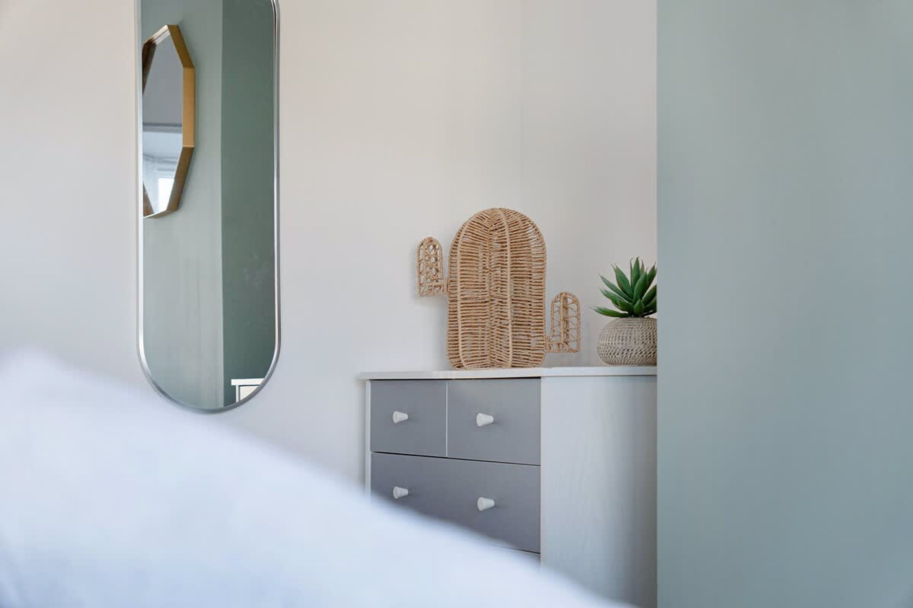 Ein Schlafzimmer mit einem Bett, einer Kommode mit Schubladen, einem Spiegel und einem Korbwäschekorb darauf.