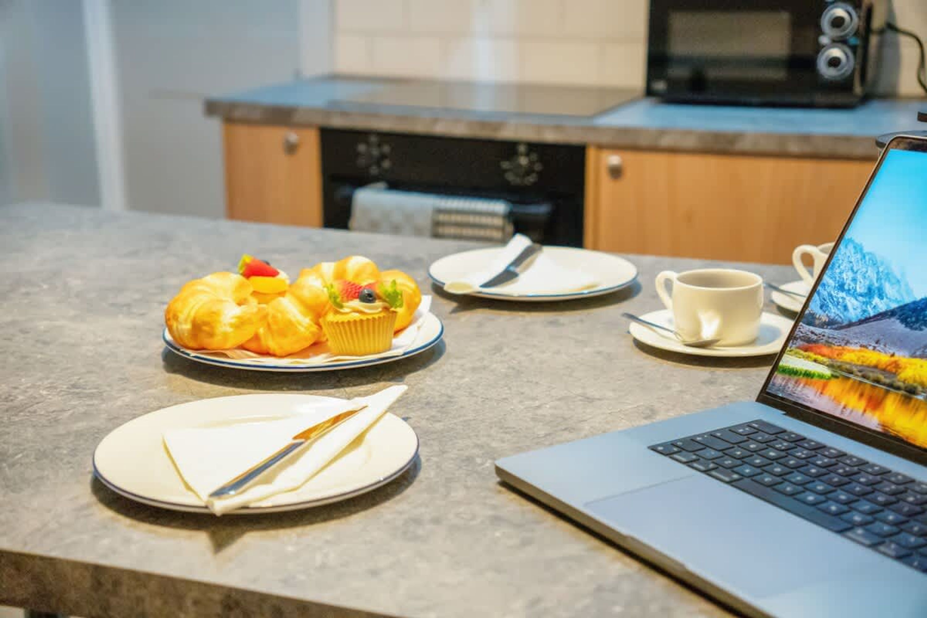 A laptop on a table with a muffin and a cup of coffee, a plate with a knife, and a microwave in the background.