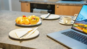 Een laptop op een tafel met een muffin en een kop koffie, een bord met een mes, en een magnetron op de achtergrond.