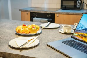 Ein Laptop auf einem Tisch zusammen mit einem Muffin und einer Kaffeetasse, ein Teller mit einem Messer und eine Mikrowelle im Hintergrund.