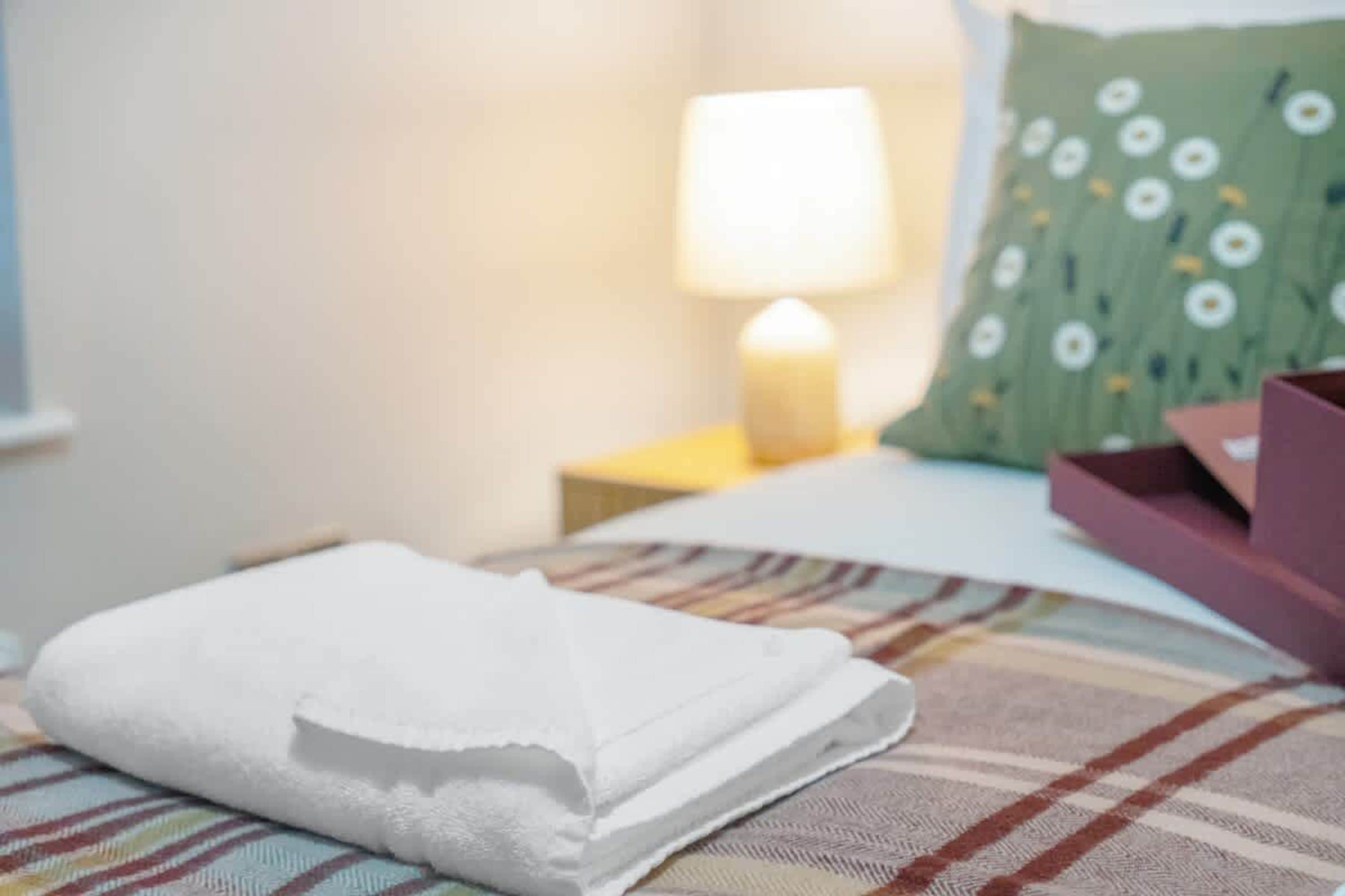 En snyggt bäddad säng med en vikt vit handduk, en grön kudde med ett prästkragsmönster och ett sängbord med en lampa.