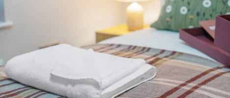 A neatly made bed with a folded white towel, a green pillow with a daisy pattern, and a bedside table with a lamp.