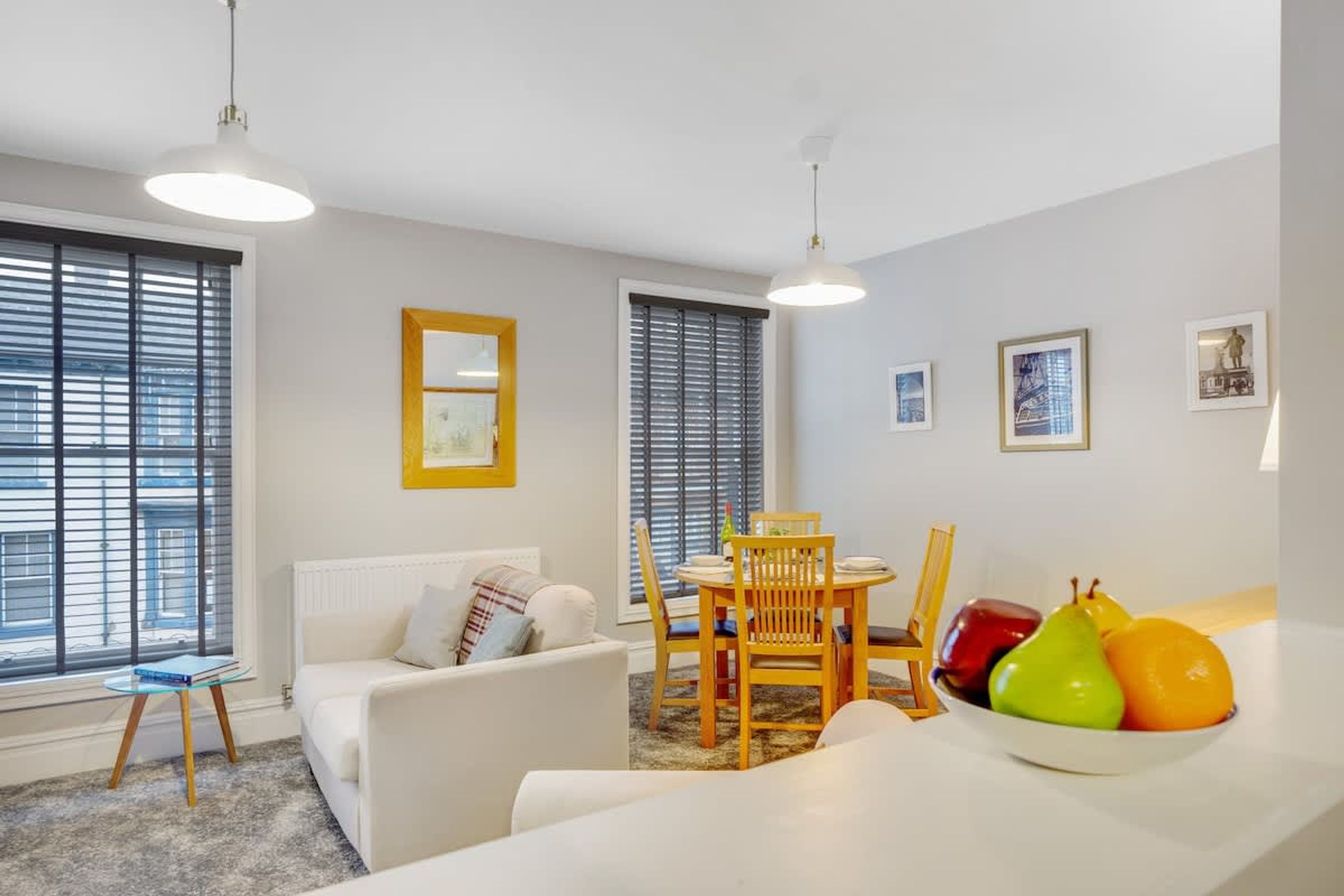 A modern living room with a white sofa, a dining table with chairs, and a fruit bowl.