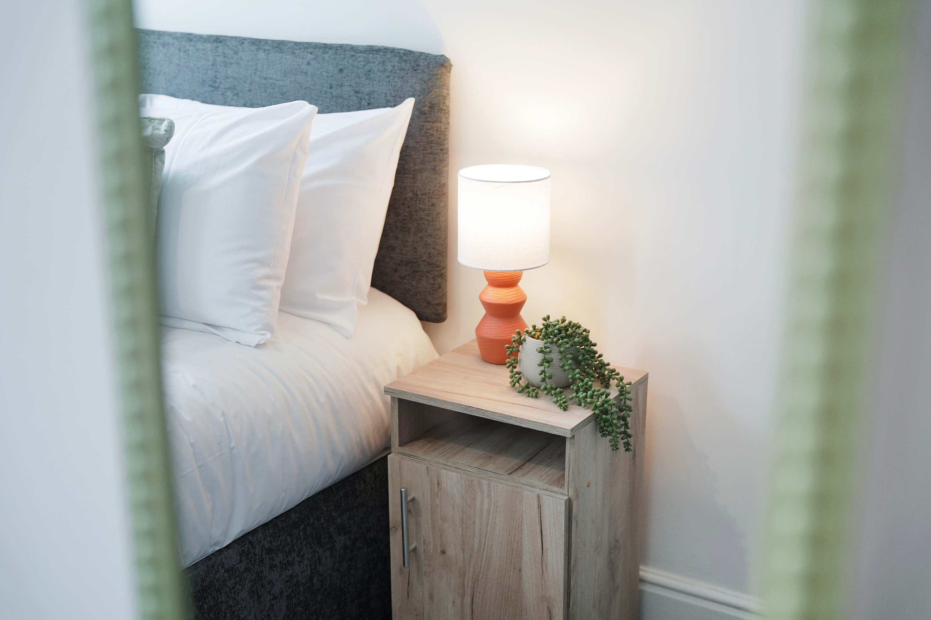 Een net opgemaakt bed met witte lakens en kussens, een nachtkastje met een lamp en een kleine potplant.