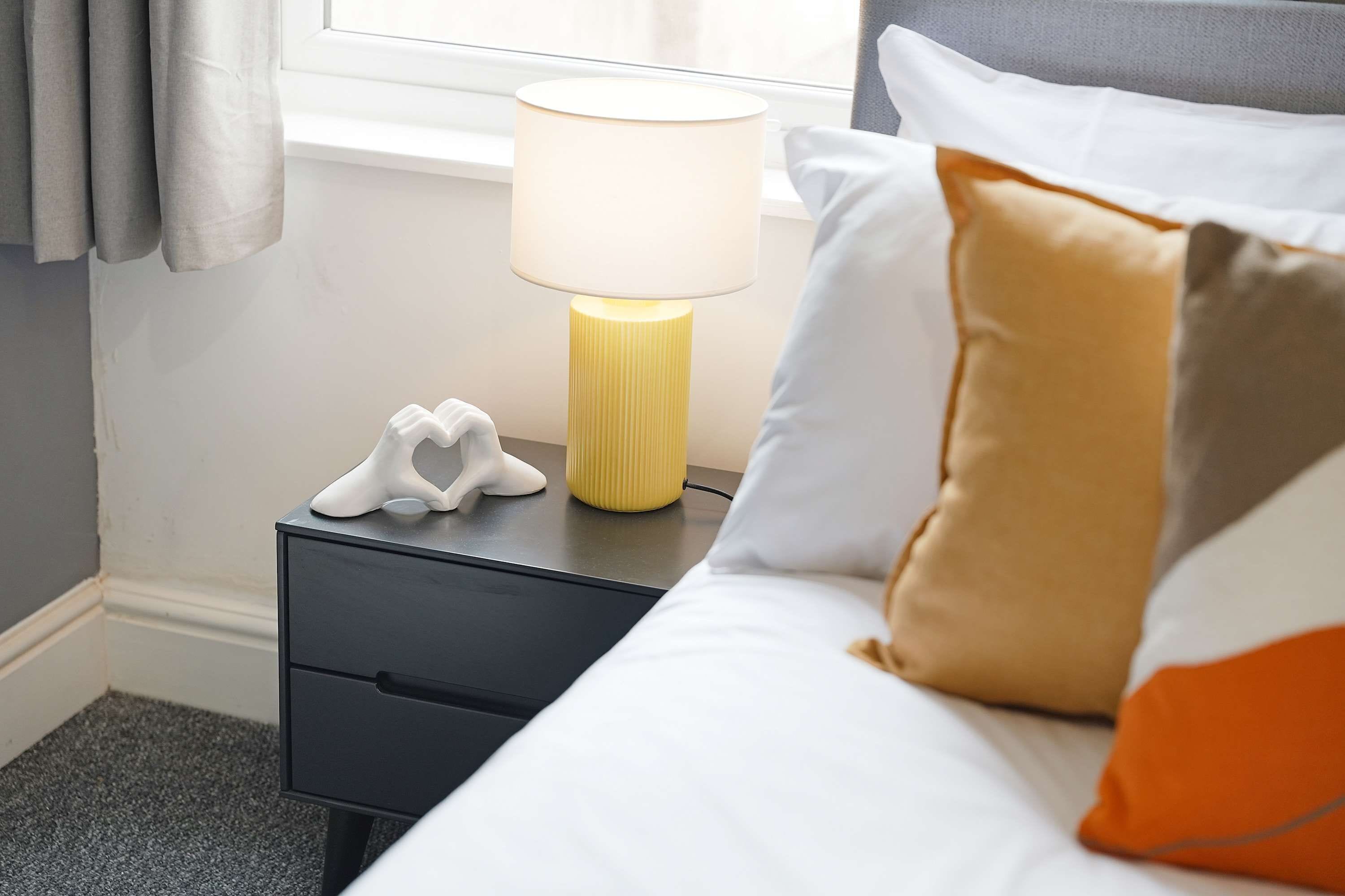 Una cama bien hecha con almohadas, una lámpara de noche y un objeto blanco pequeño en una mesita.
