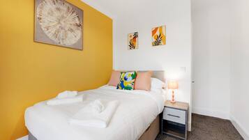 Una habitación de hotel con cama, mesita de noche, lámpara y una obra de arte en la pared.