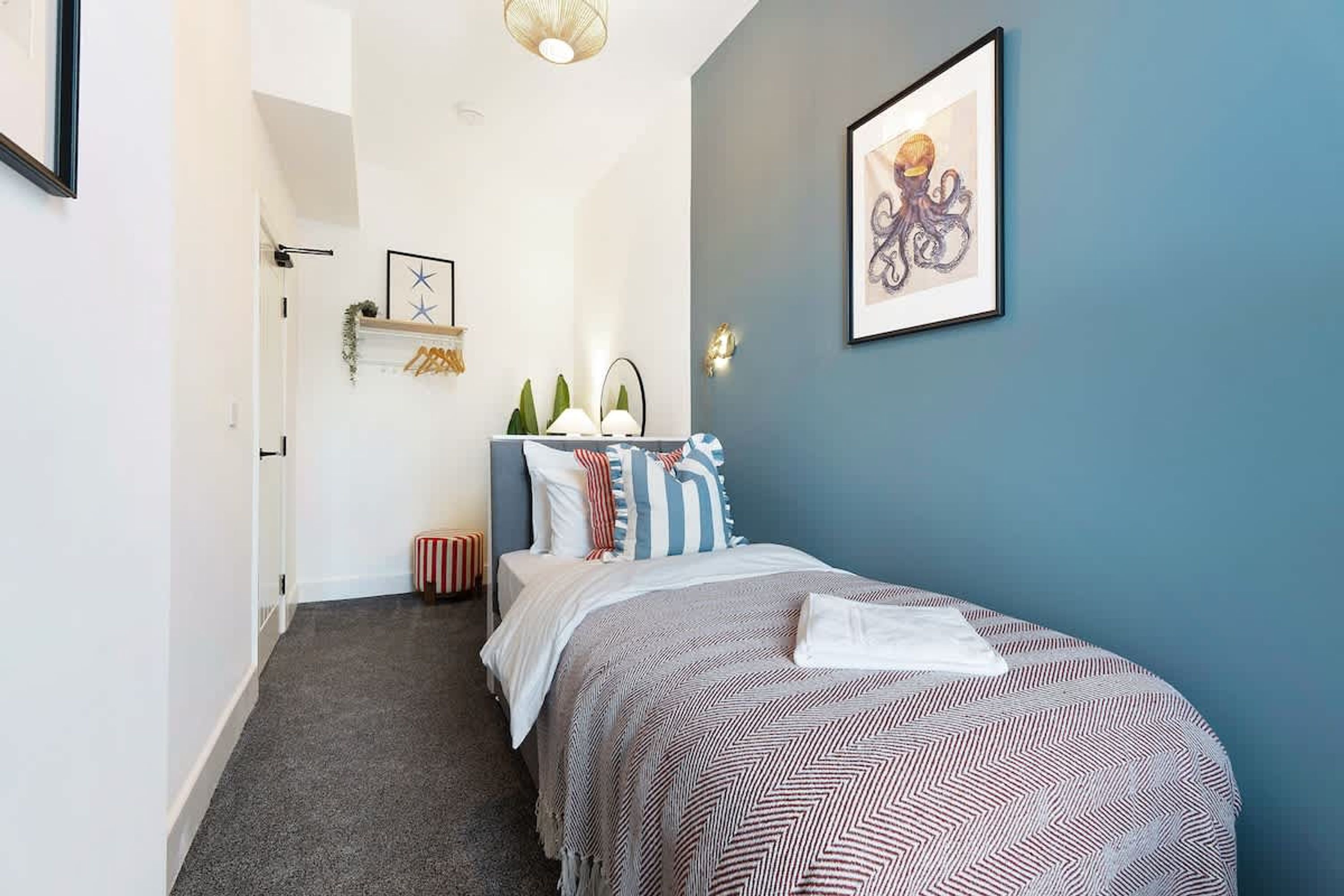 Un dormitorio con una cama, una mesita de noche, una lámpara de pared, un cuadro enmarcado y una puerta que da a otra habitación.