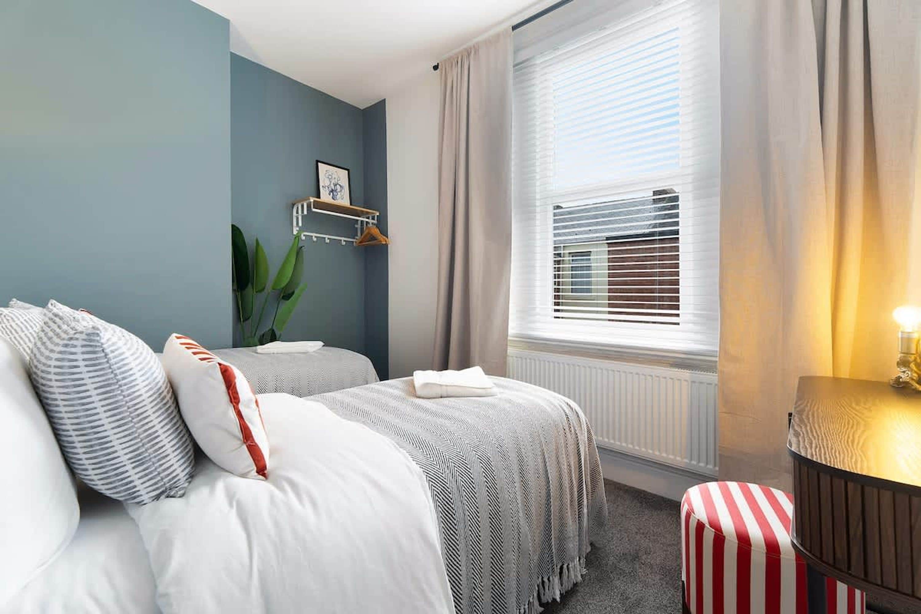 Quarto de hotel com duas camas, um radiador, uma janela com persianas, uma mesa de cabeceira e uma planta.