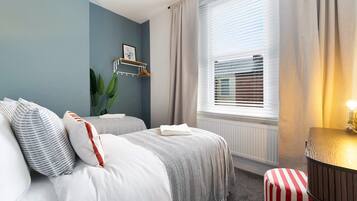 Quarto de hotel com duas camas, um radiador, uma janela com persianas, uma mesa de cabeceira e uma planta.