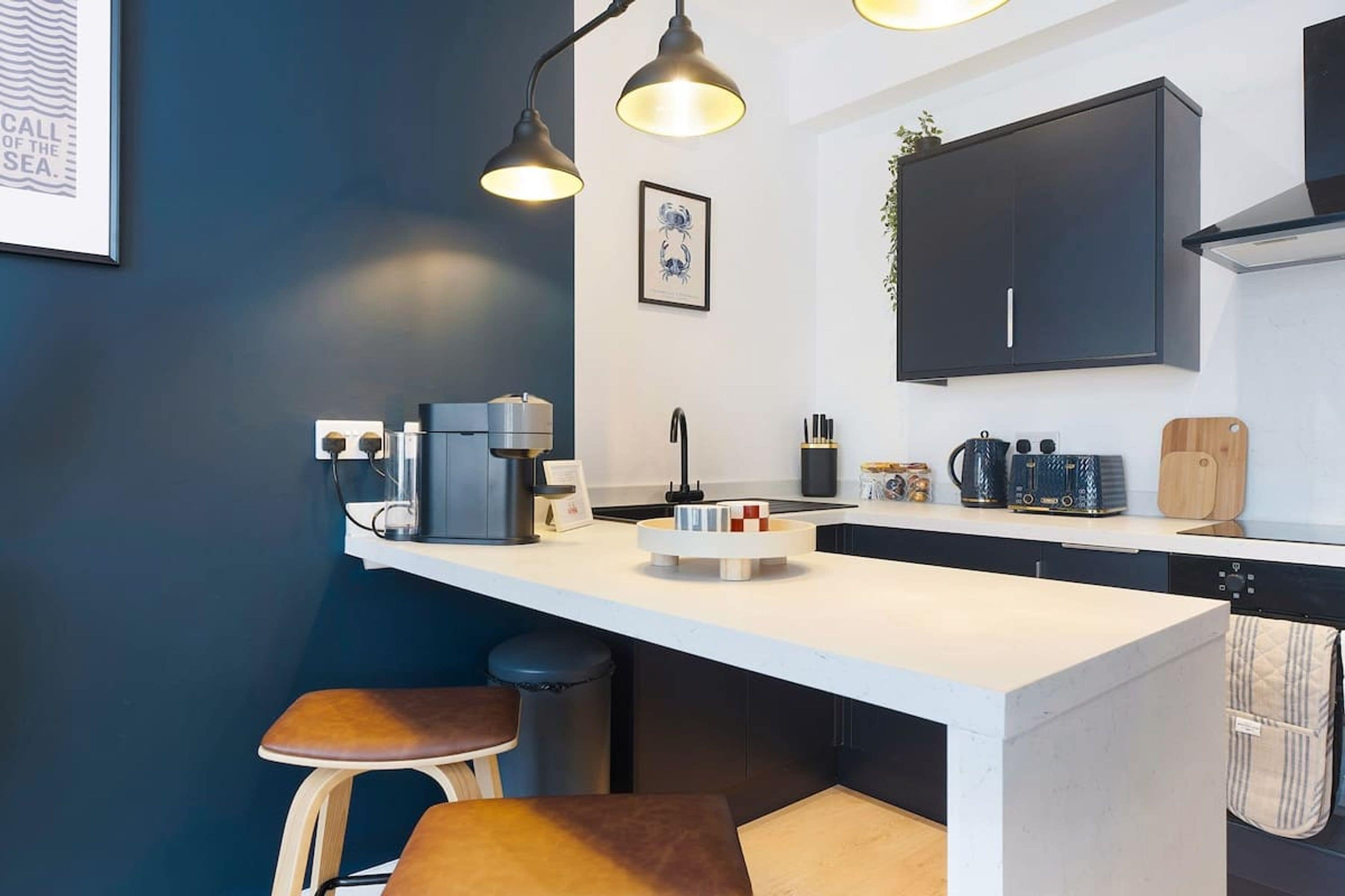 A modern kitchen with a white countertop, two bar stools, a coffee machine, a kettle, and a black cabinet.