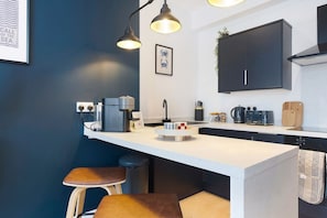 Eine moderne Küche mit weißer Arbeitsplatte, zwei Barhocker, einer Kaffeemaschine, einem Wasserkocher und einem schwarzen Schrank.