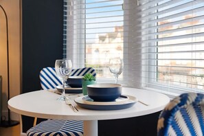Een eettafel met een blauwe schaal en twee wijnglazen, voor een raam met jaloezieën.