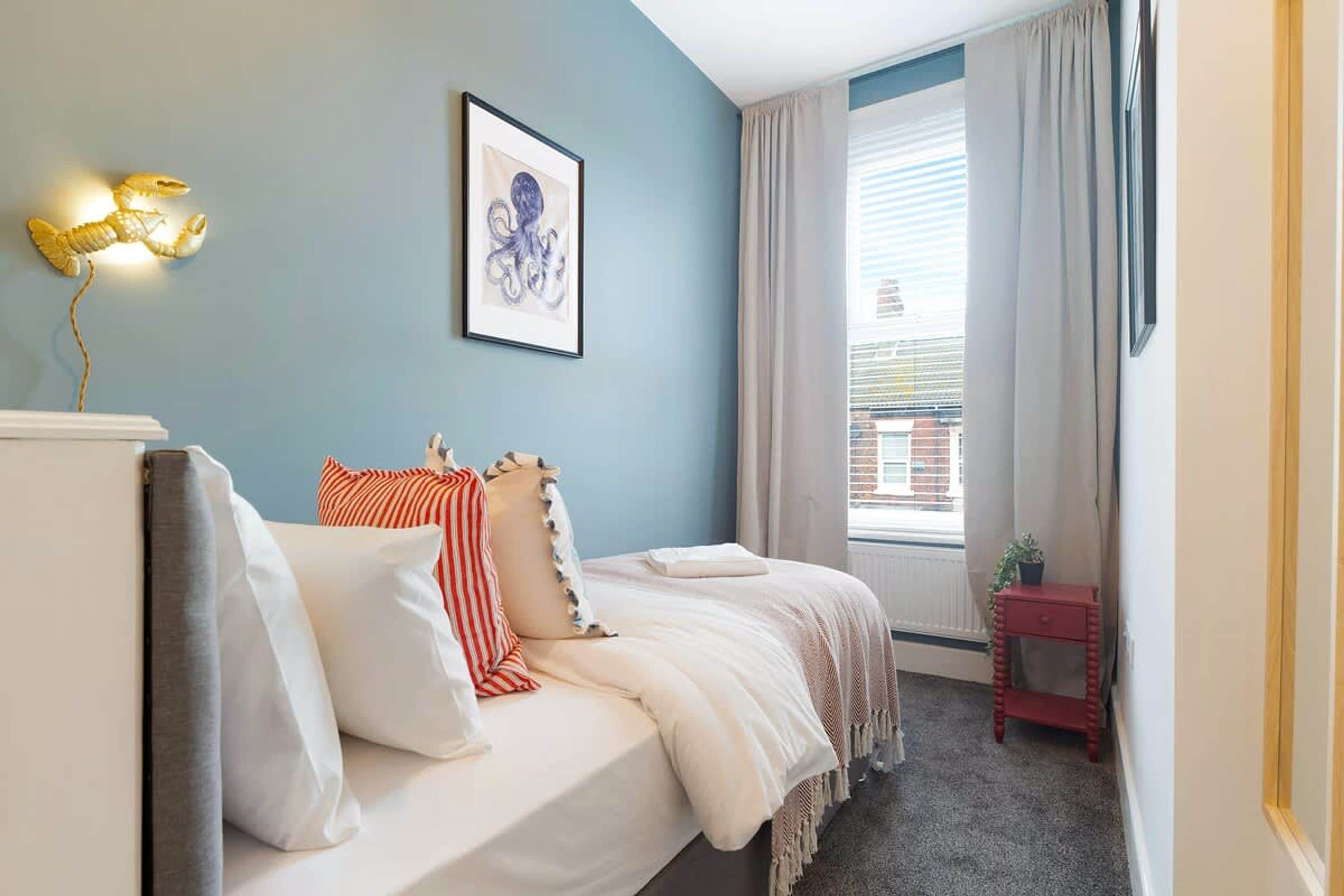 Un dormitorio con cama, almohadas, mesita de noche, lámpara, ventana con cortinas e imagen enmarcada en la pared.