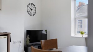 Ein modernes Wohnzimmer mit Flachbildfernseher, einem Esstisch, einem Sessel, einer Uhr und einem Fenster mit Blick auf Gebäude.