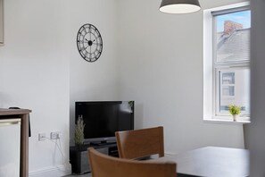 Ein modernes Wohnzimmer mit Flachbildfernseher, einem Esstisch, einem Sessel, einer Uhr und einem Fenster mit Blick auf Gebäude.