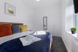 Una camera d'albergo con un letto, cuscini, un comodino, uno specchio, una finestra con una pianta e un termosifone.