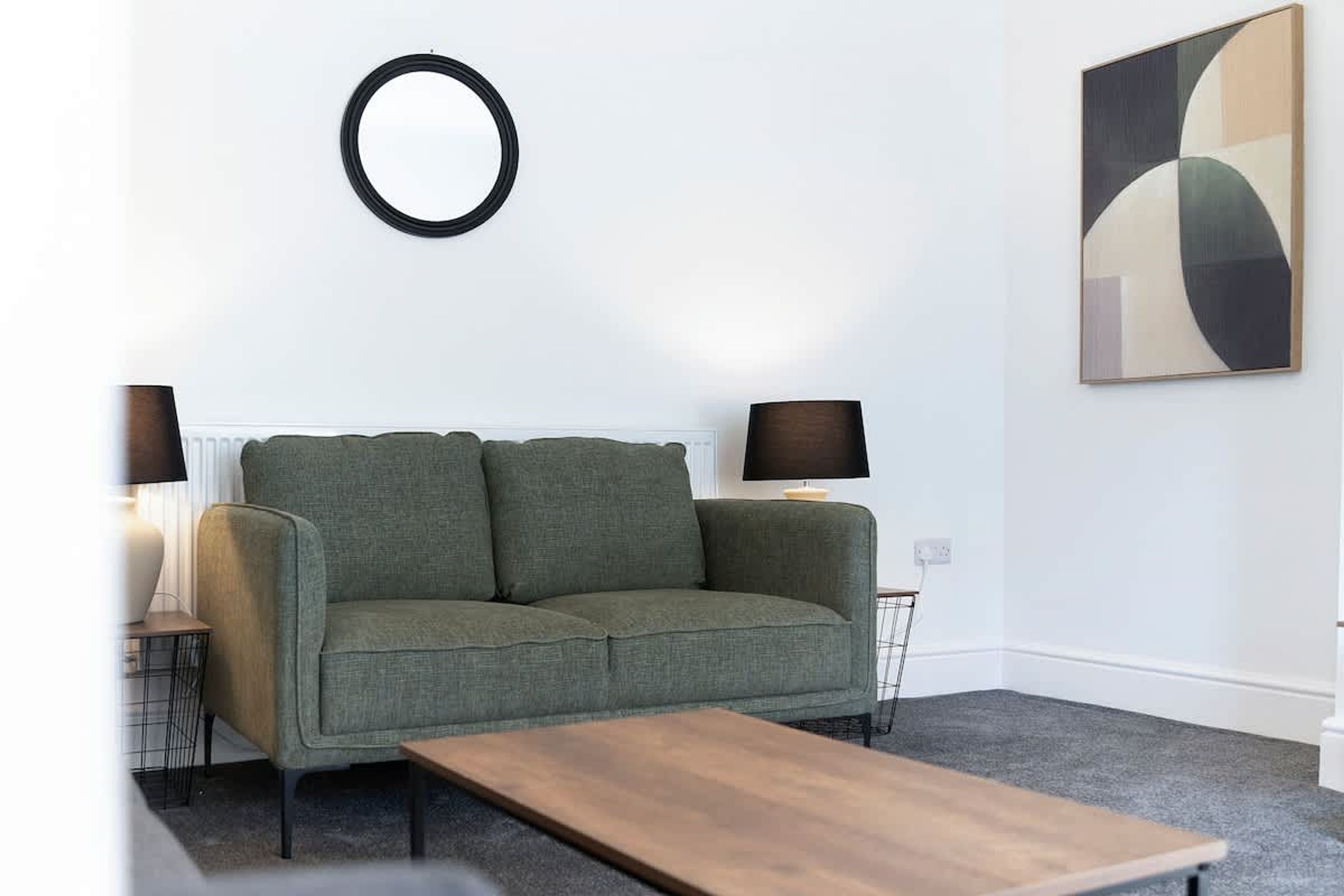 A living room with a green sofa, two side tables with lamps, a coffee table, a wall-mounted circular mirror, and a large abstract painting.