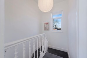 Un escalier avec une rampe blanche, une petite fenêtre donnant sur les arbres et un luminaire fixé au mur.