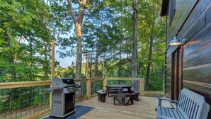 Eine Holzterrasse mit Grill, Picknicktisch und Bank, umgeben von Bäumen.