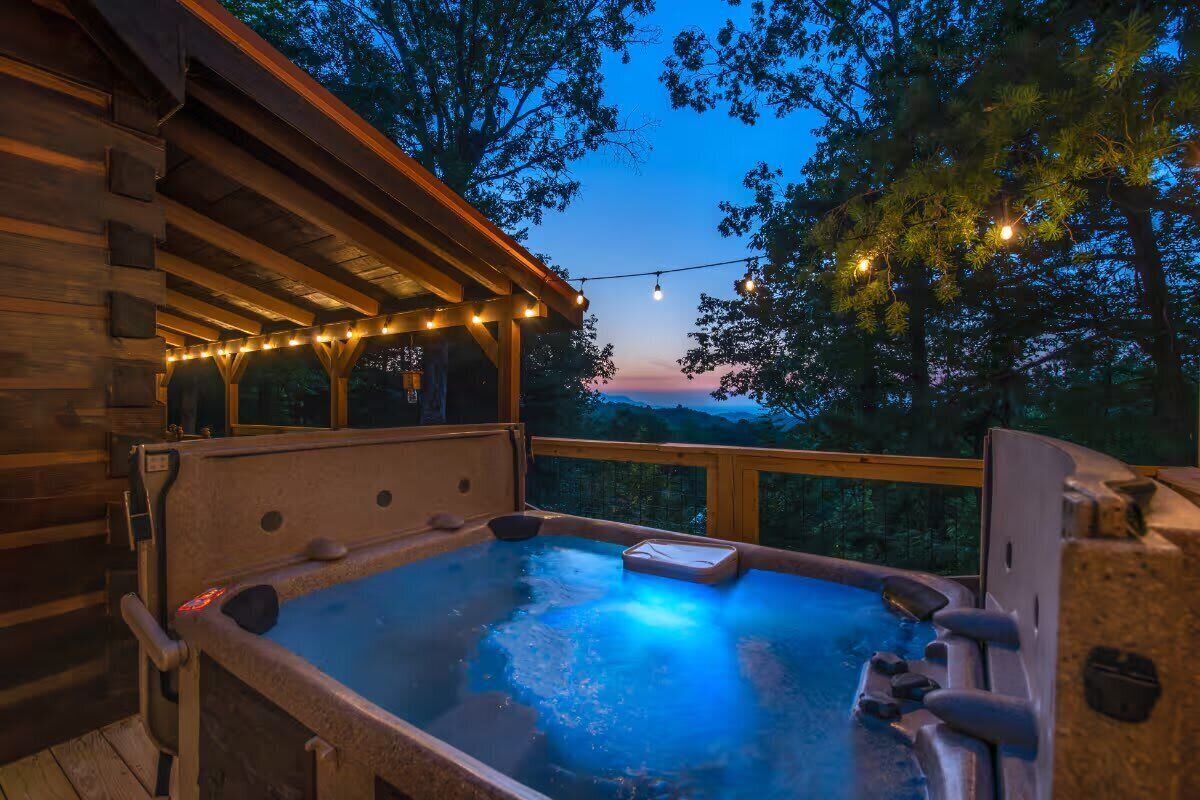 A hot tub with a wooden deck and string lights, surrounded by trees.