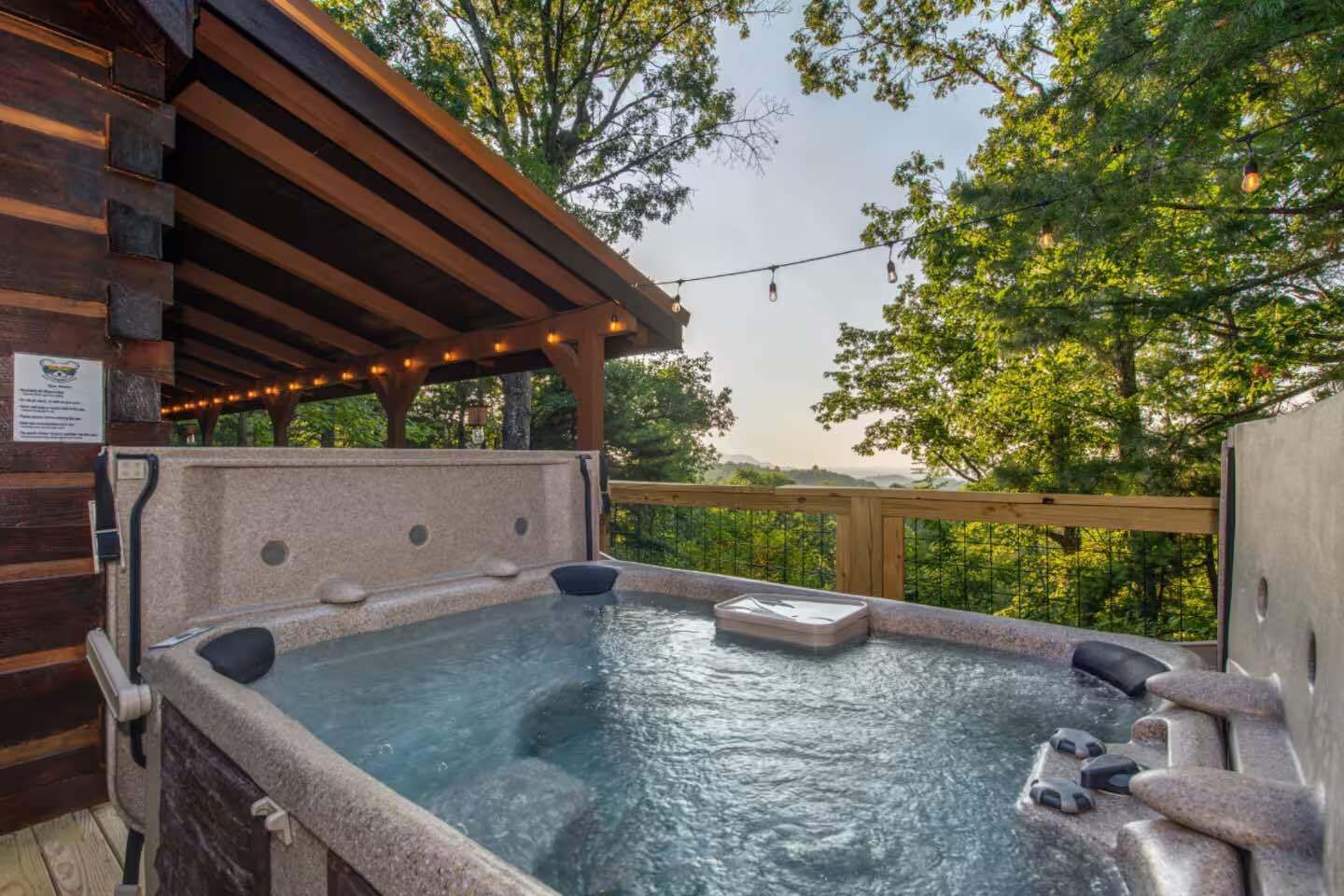 A hot tub with a wooden deck and string lights, surrounded by trees.