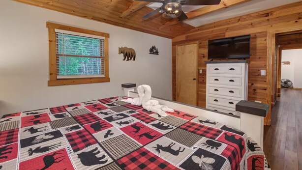Ein Schlafzimmer mit Holzbalkendecke, Deckenventilator, Bett mit kariertem Überwurf, Kommode und Fenster mit Jalousien.