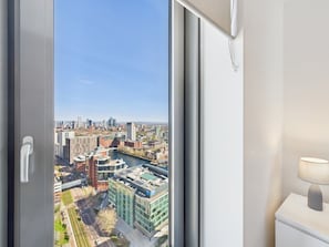 Una habitación con un ventanal que ofrece vistas a un paisaje urbano, una mesita de noche blanca con una lámpara, y una puerta con manija.