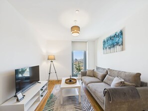 A modern living room with a flat-screen TV, a white coffee table, a floor lamp, a sofa, and a city view through a large window. - New! 1BR in Media City (Salford)