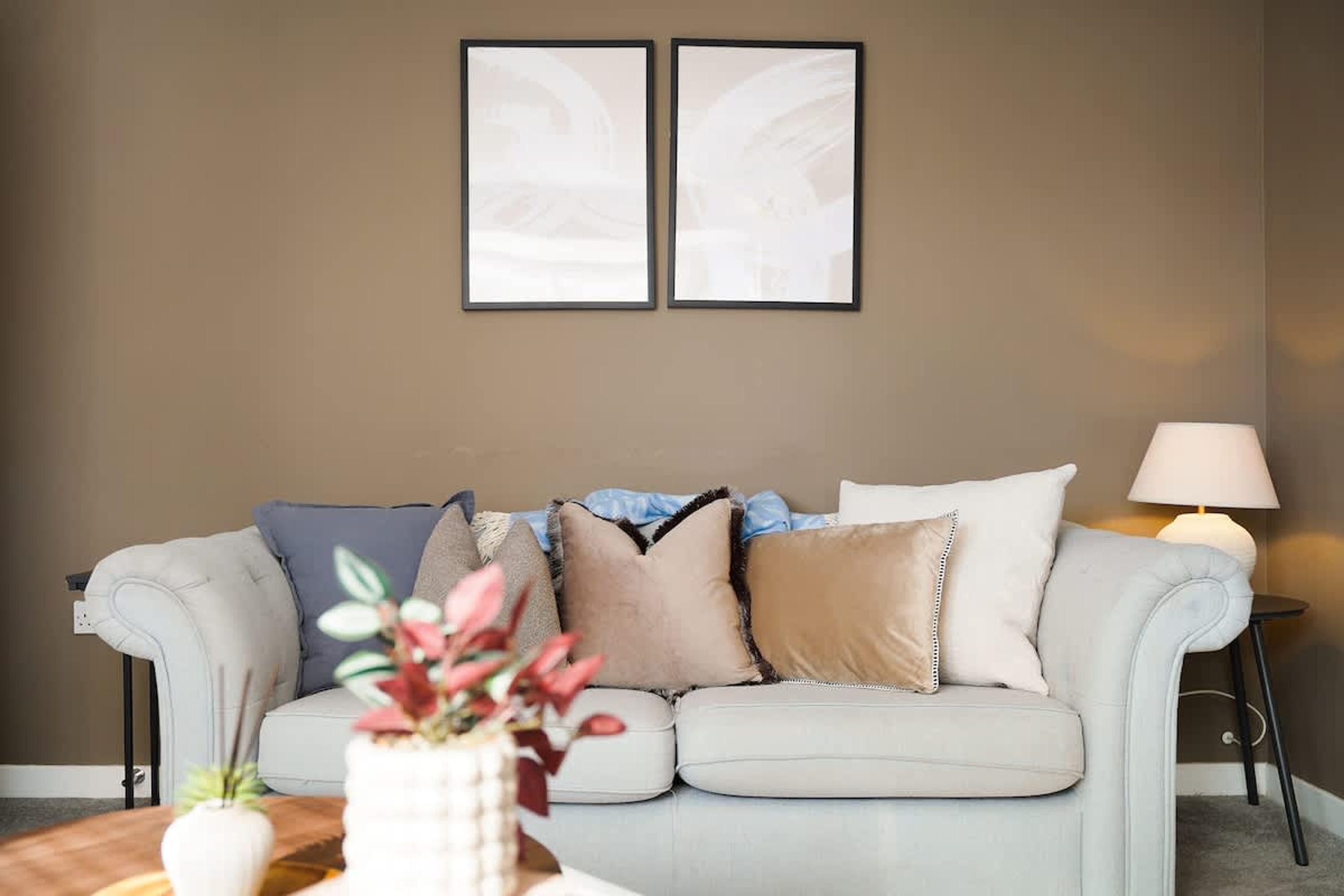A living room with a light-colored sofa, a side table with a lamp, and two framed pictures on the wall.