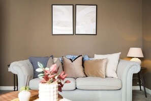 Ein Wohnzimmer mit einem hellen Sofa, einem Beistelltisch mit Lampe und zwei gerahmten Bildern an der Wand.