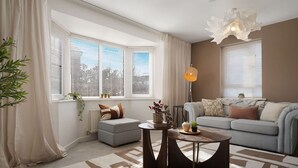Una sala de estar moderna con sofá, mesa de centro y lámpara. Hay ventanas con persianas, un radiador y una planta.