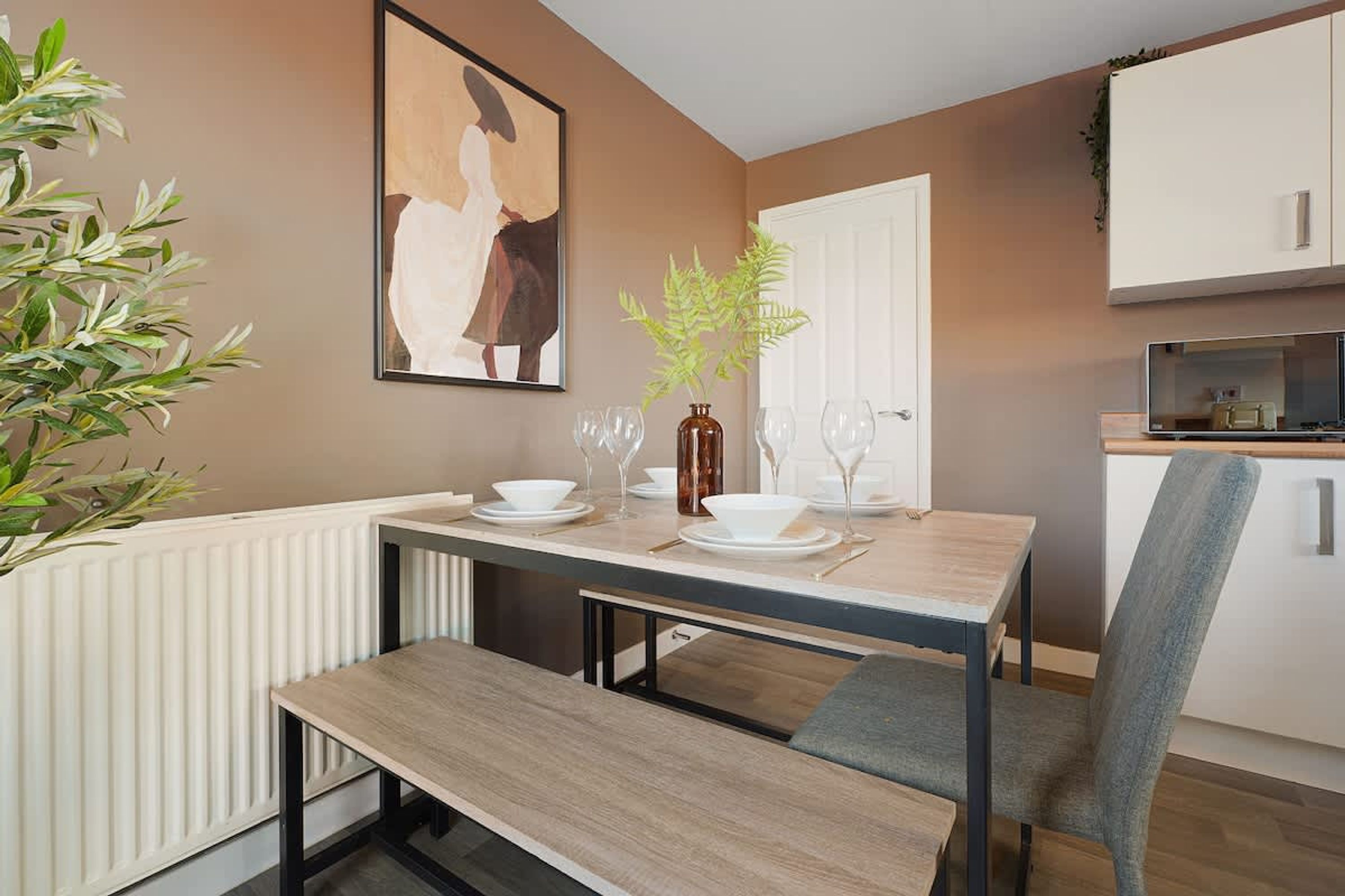 Un comedor con una mesa dispuesta para dos personas, una banca, una planta en maceta y un cuadro en la pared.