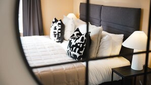 Una cama con colcha blanca y almohadas estampadas en blanco y negro, cabecera oscura y mesita de noche con lámpara y caja de pañuelos.
