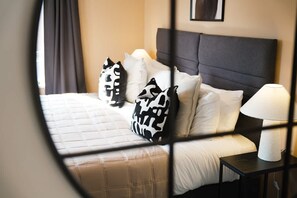 Ein Bett mit weißer Steppdecke, Kissen in Schwarzweißmuster, einem dunklen Kopfteil und einem Nachttisch mit Lampe und Taschentuchbox.