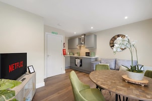 Un salón moderno con televisor de pantalla plana, una mesa de comedor con sillas, una zona de cocina con armarios y un microondas, y una orquídea en maceta en la pared.