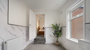Un pasillo con pared de mármol, un radiador, una planta en maceta y una ventana con vista a un muro de ladrillo.