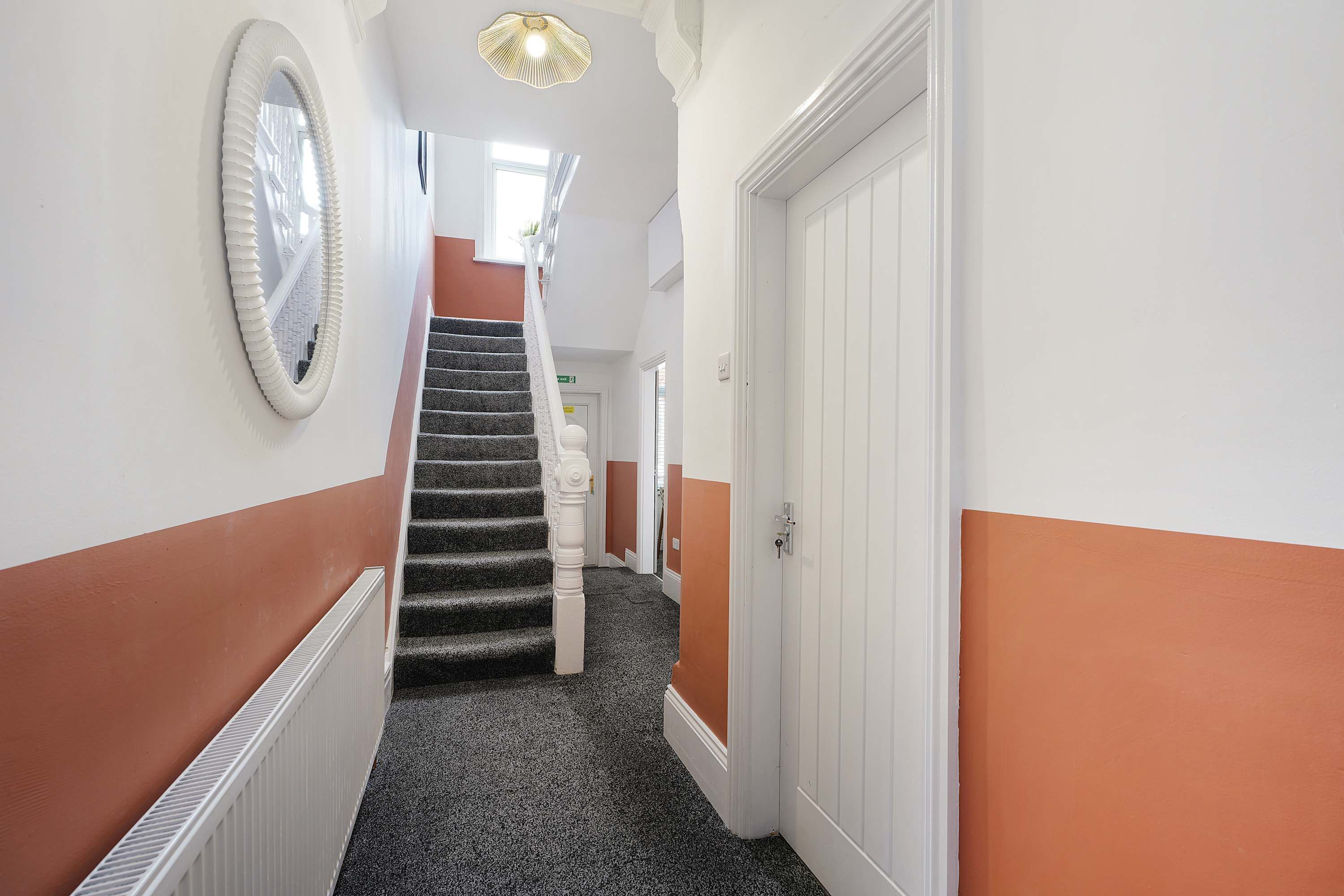 A hallway with a carpeted staircase, white walls, and a large mirror on the left.
