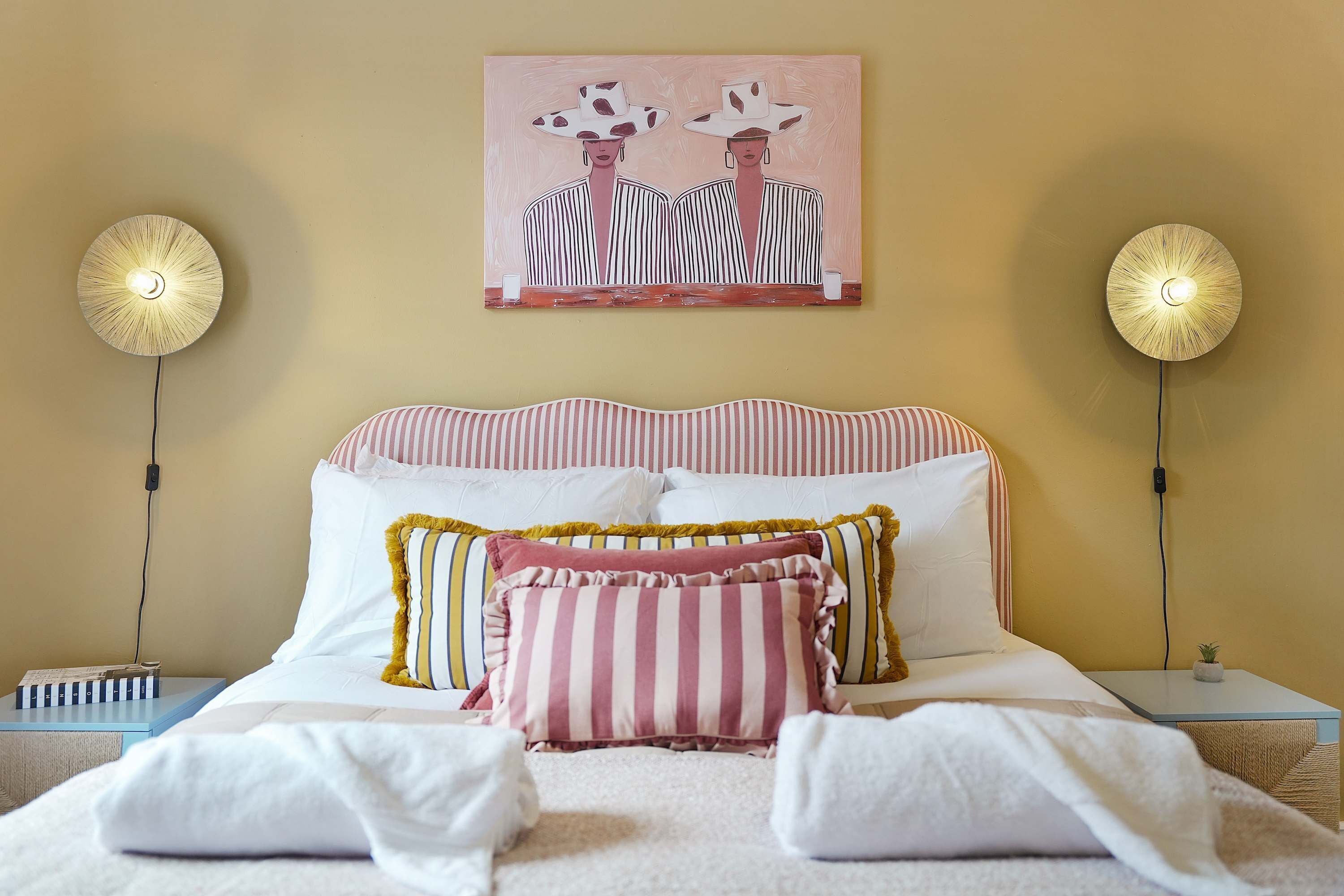 A hotel room with a bed, striped pillows, a painting above the bed, and two wall-mounted lamps.