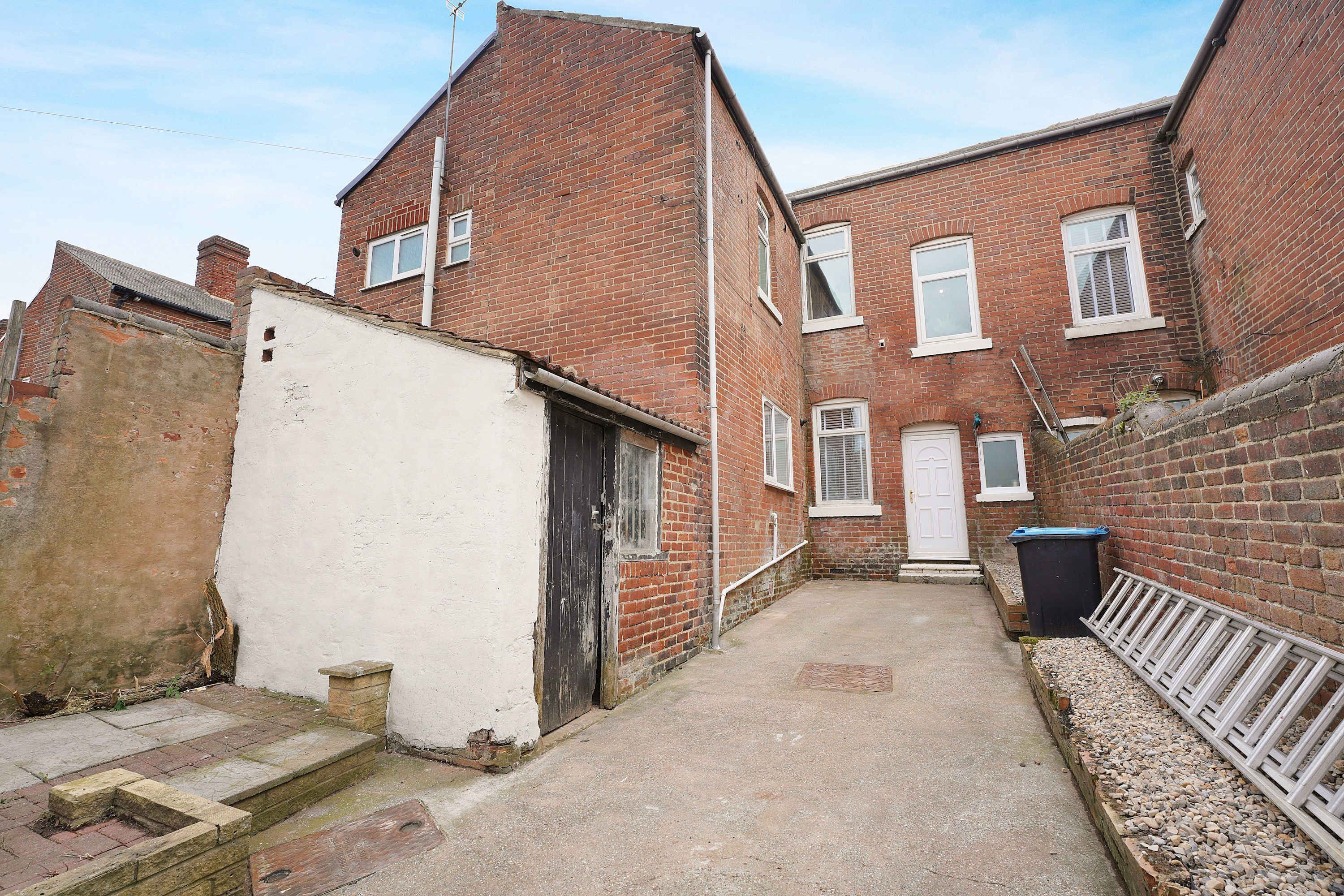 A narrow alleyway between two brick buildings with a white door and a black door, a white fence on the right, and a black trash bin.